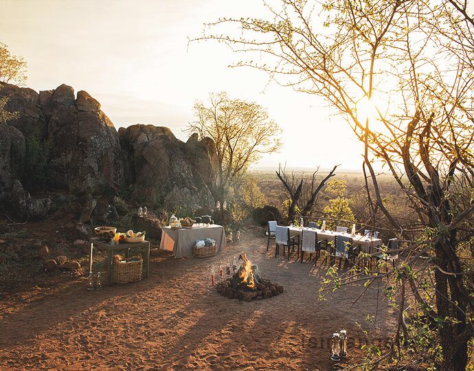 A long table laid for dinner beside a campfire below a rocky outcrop at sunset