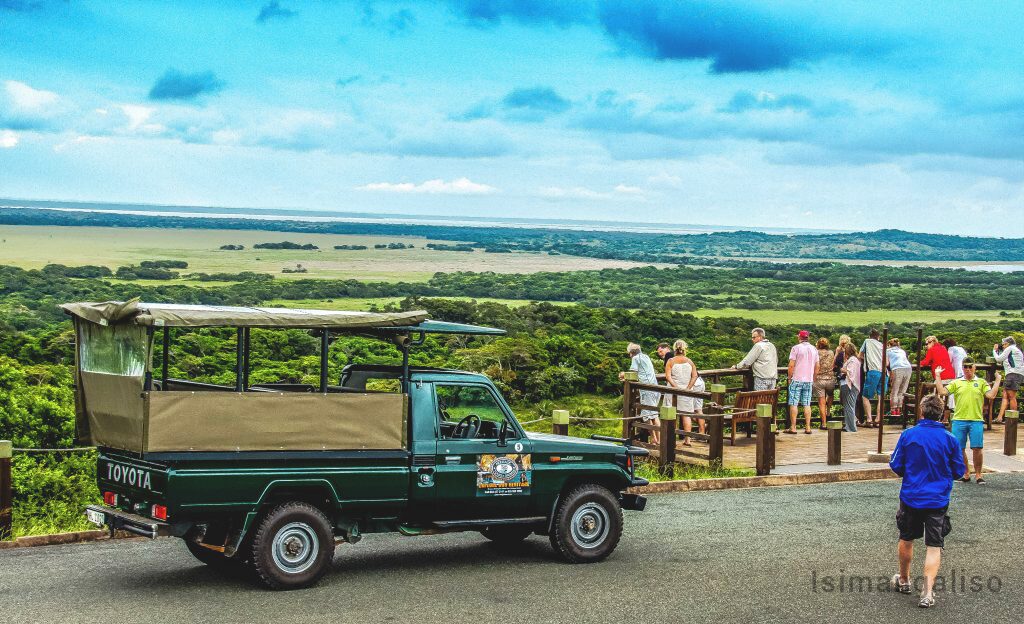 Visitors at a viewpoint railing above coastal forest and lake, beside an open-sided game-drive vehicle